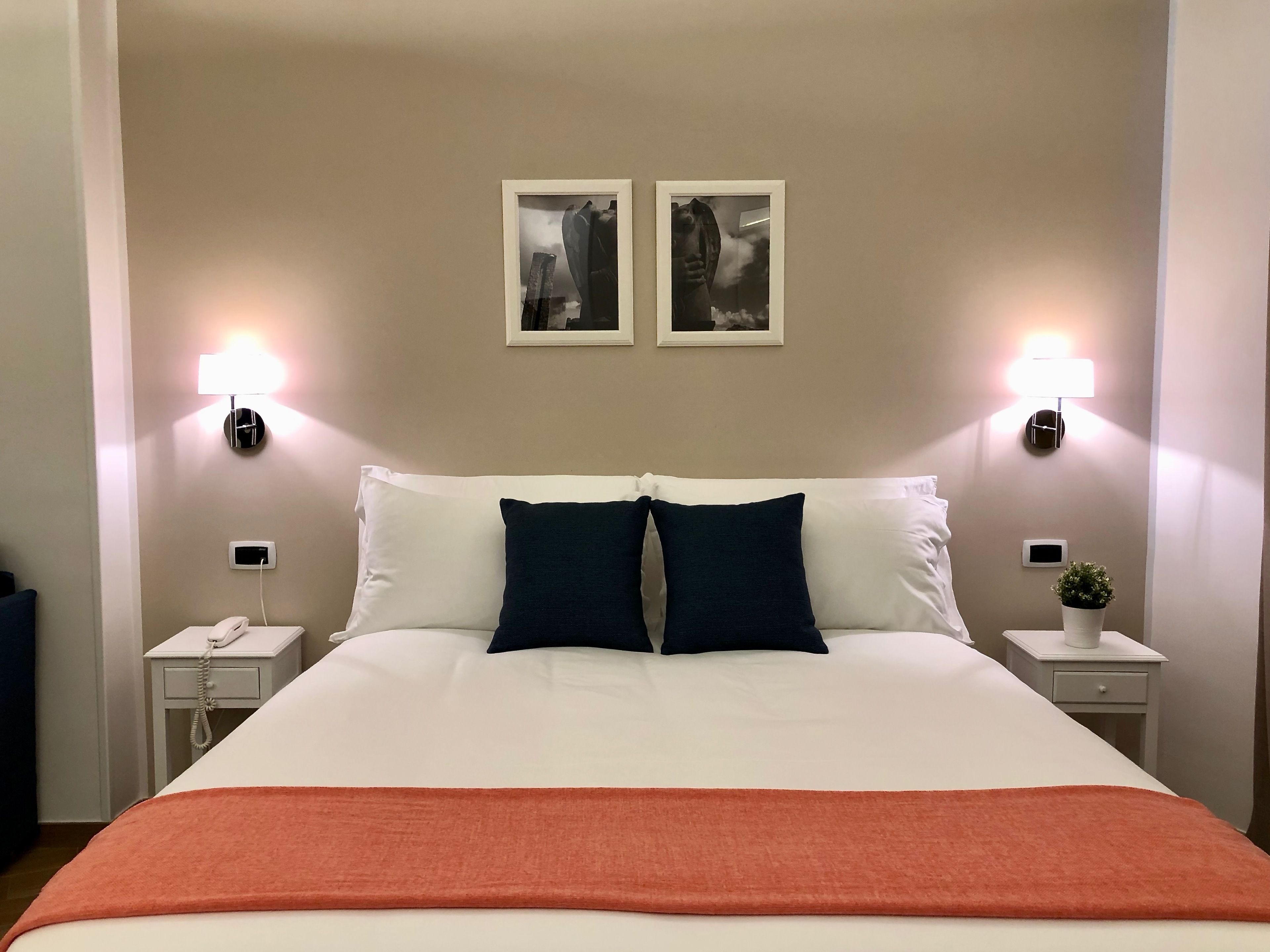 A neatly made bed with white and dark blue pillows, two wall-mounted lamps, and a small potted plant on a bedside table.