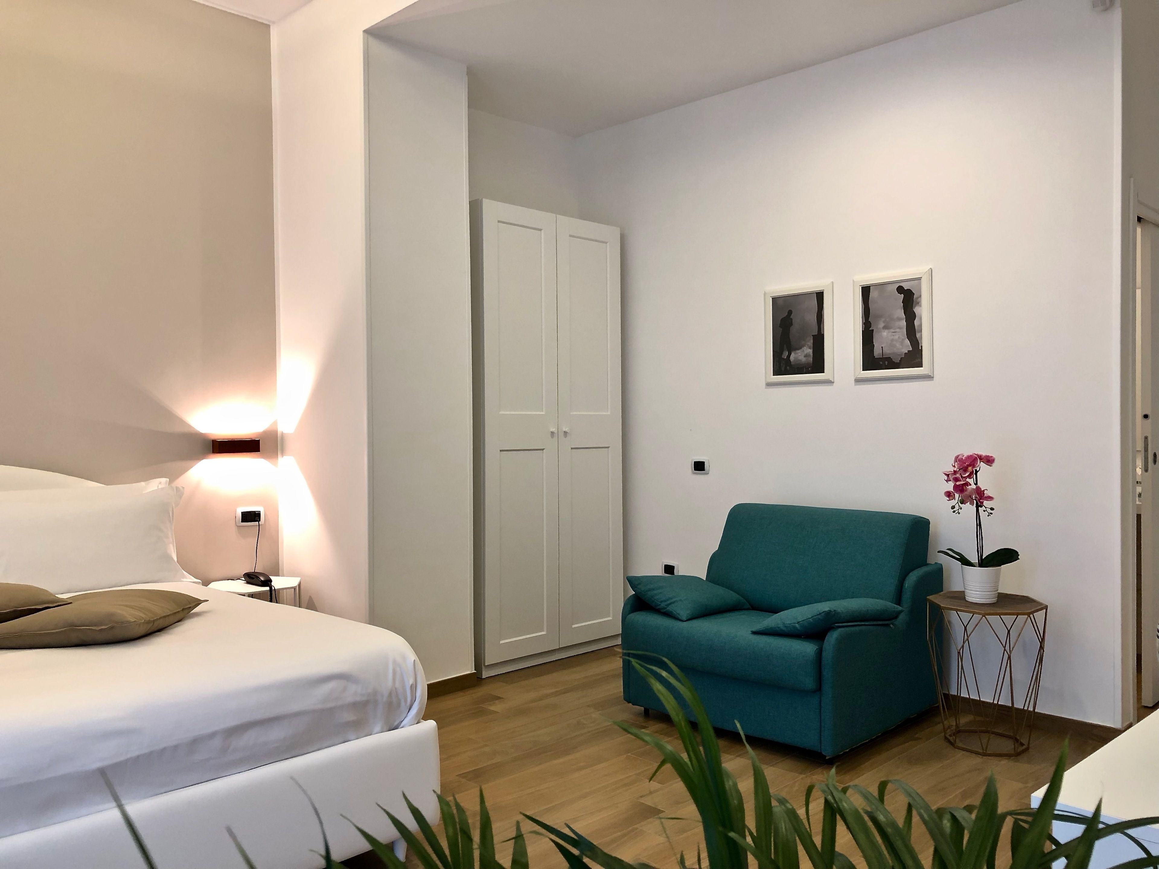 Ein Schlafzimmer mit einem Bett, einem türkisblauen Sessel, einem kleinen Beistelltisch mit Orchidee im Topf und einem weißen Kleiderschrank.