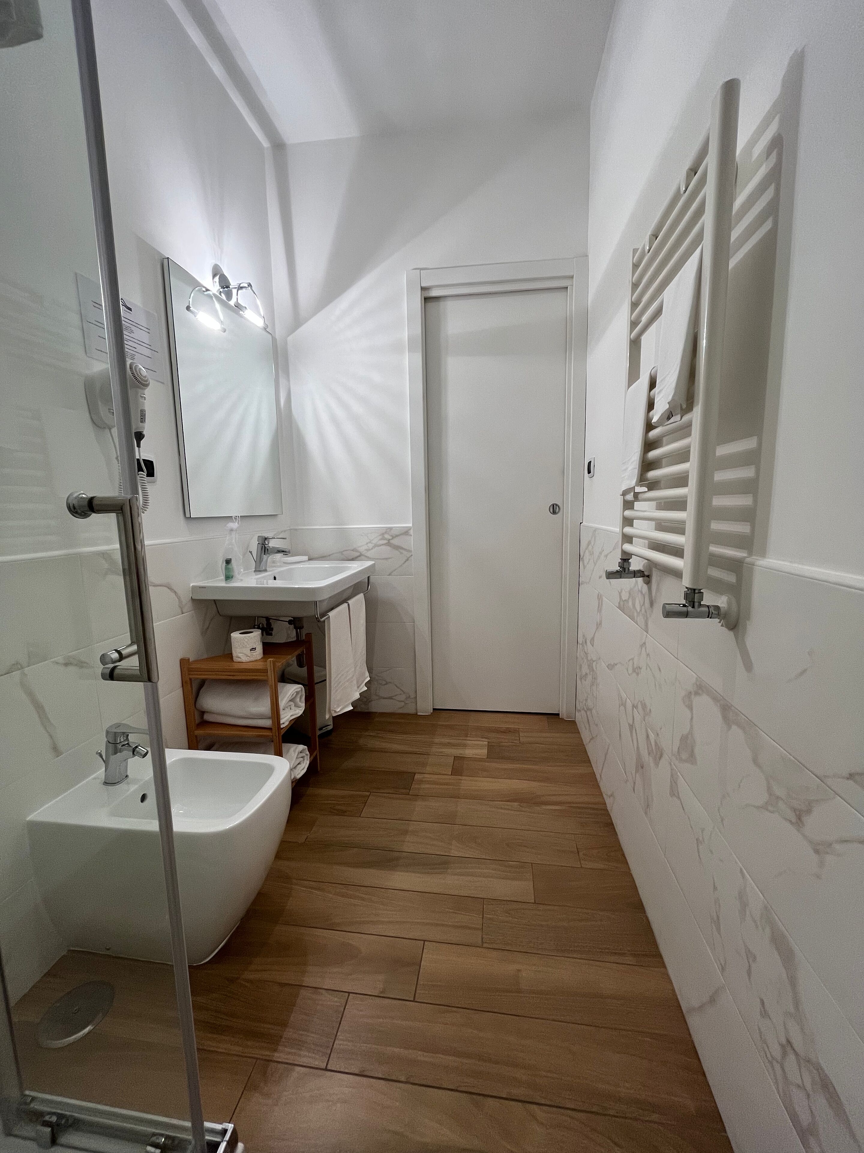 A narrow bathroom with a sink, bidet, and mirror. There is a towel rack on the wall and a wooden shelf with towels.
