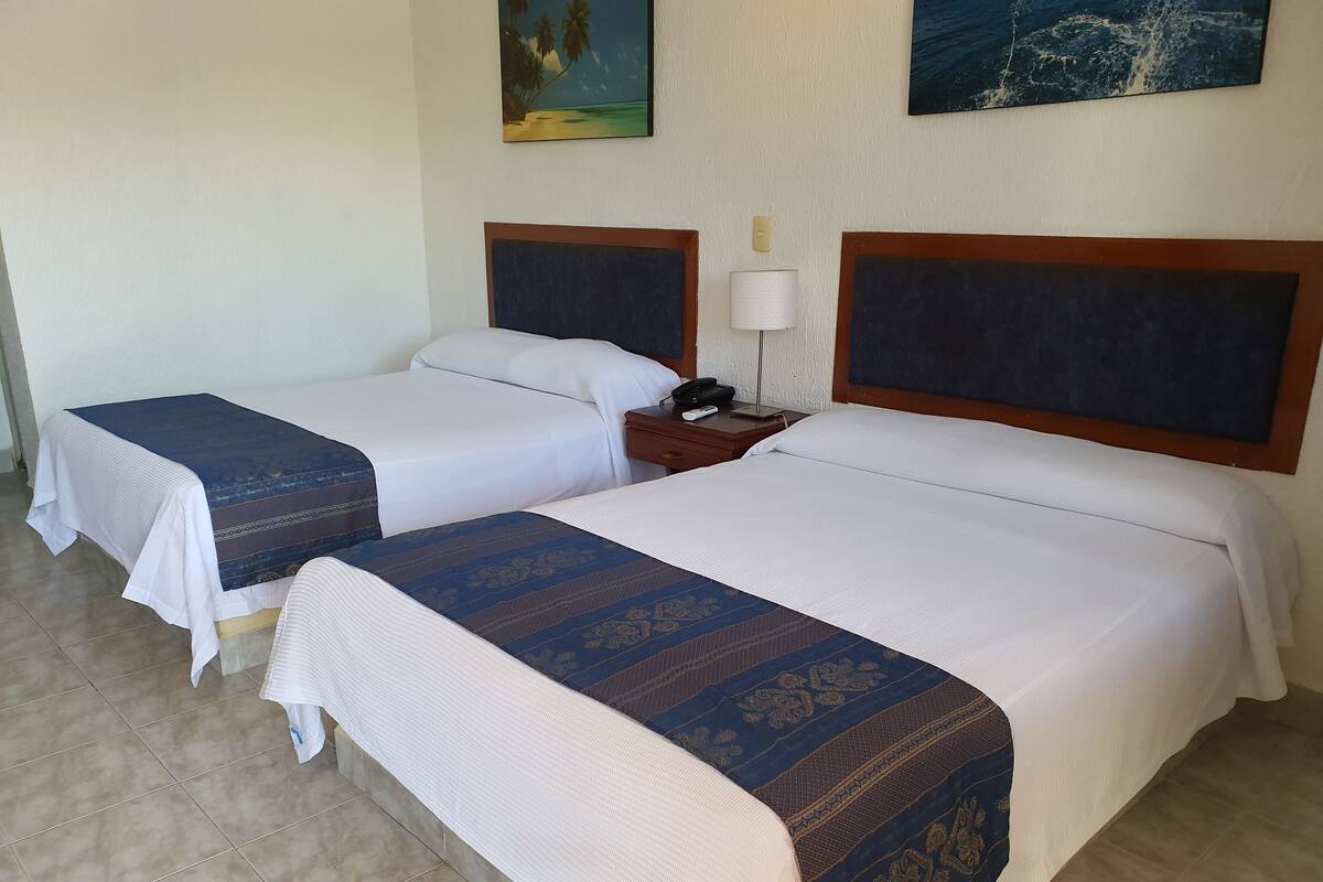 Habitació d'hotel amb dos llits, cadascun amb una mantella blanca i un petit davant blau estampat. Hi ha una tauleta de nit amb una làmpada i un telèfon entre els llits.
