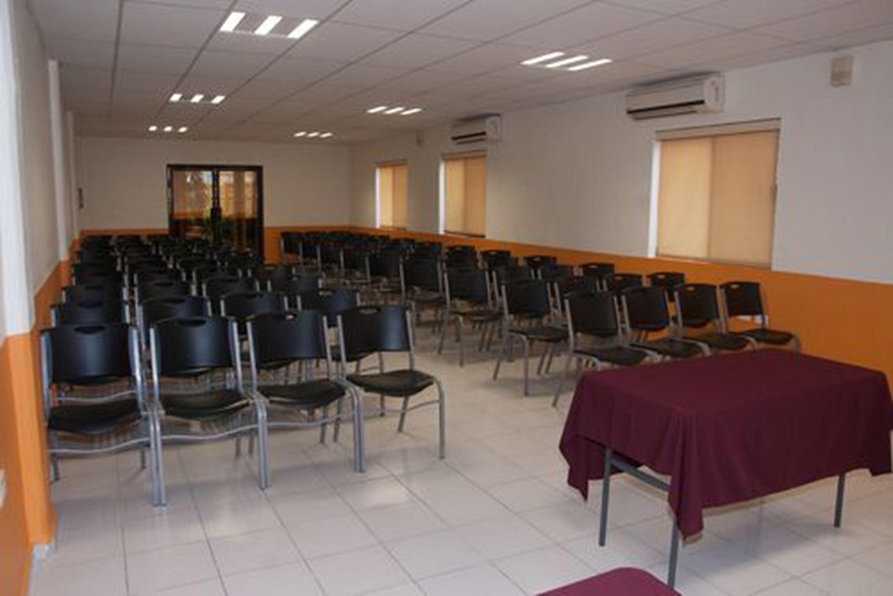 Un salón de conferencias con filas de sillas, una mesa con mantel y un equipo de aire acondicionado en la pared.