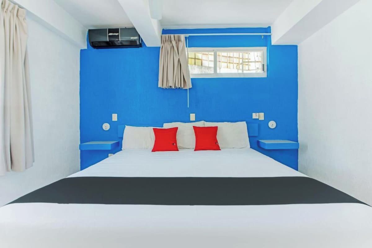 Une chambre d’hôtel avec un grand lit, des murs d’un bleu vif, une fenêtre avec des rideaux et un climatiseur au plafond.