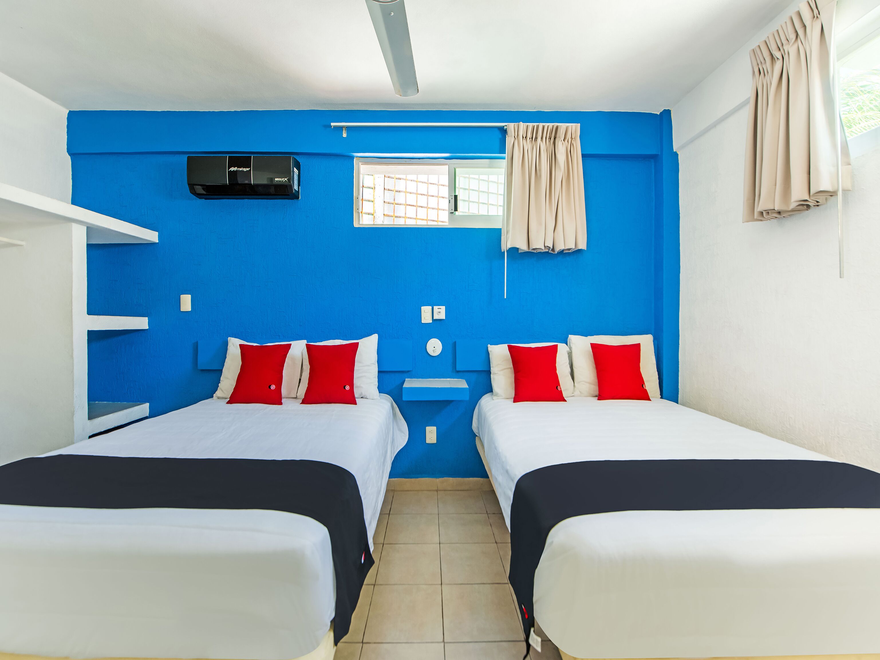 Ein Zimmer mit zwei Betten, jeweils mit rot-weißer Bettwäsche, einem Fenster mit beigen Vorhängen und einem Deckenventilator.