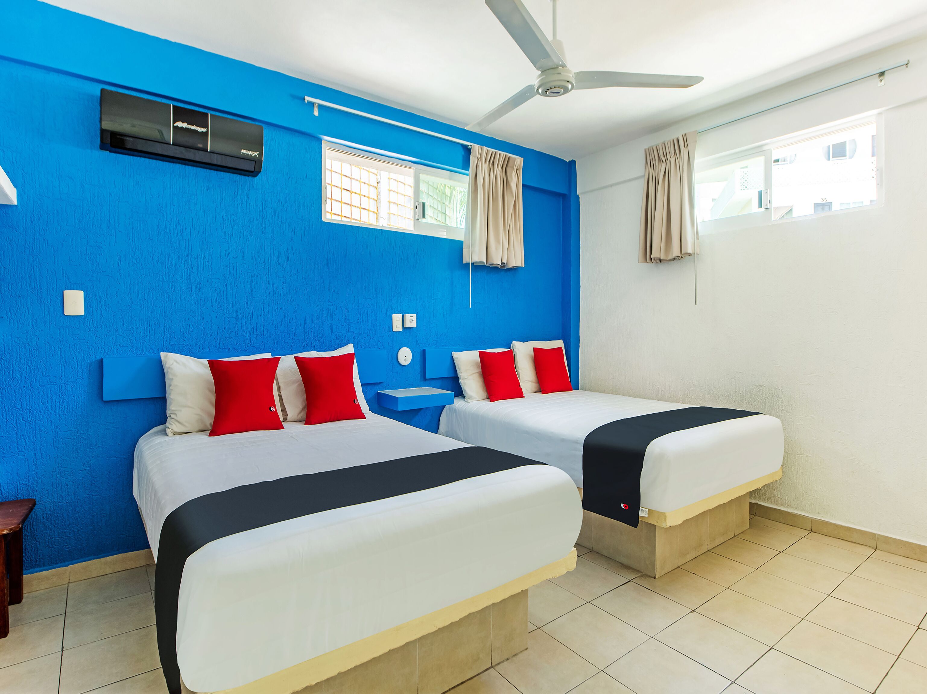 A hotel room with two beds, a ceiling fan, an air conditioning unit, and a window with curtains.