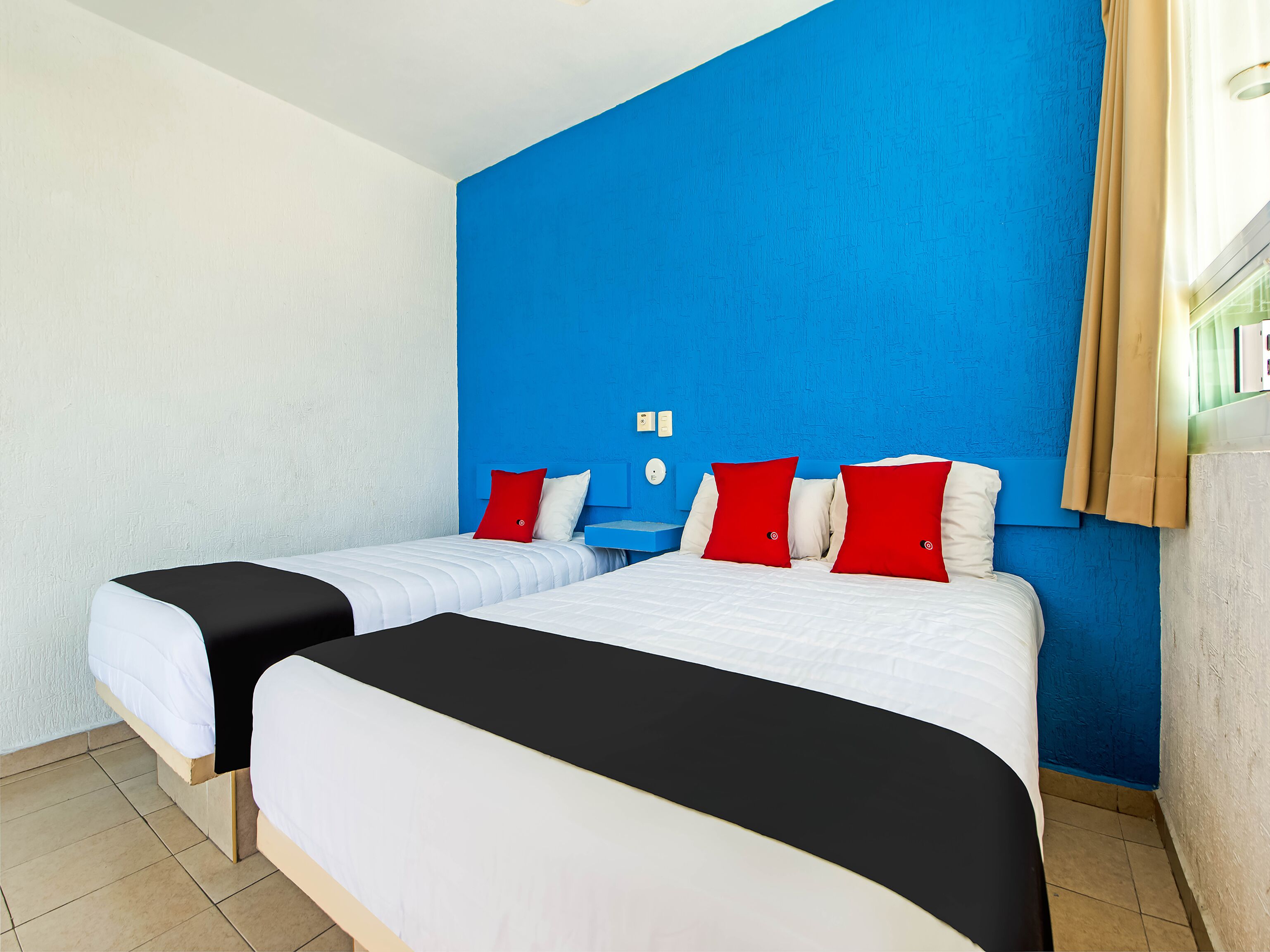 Une chambre d’hôtel avec deux lits, chacun doté de draps blancs et d’une tête de lit noire. La chambre est agrémentée d’un mur d’accent bleu et d’une fenêtre avec des rideaux de couleur beige.