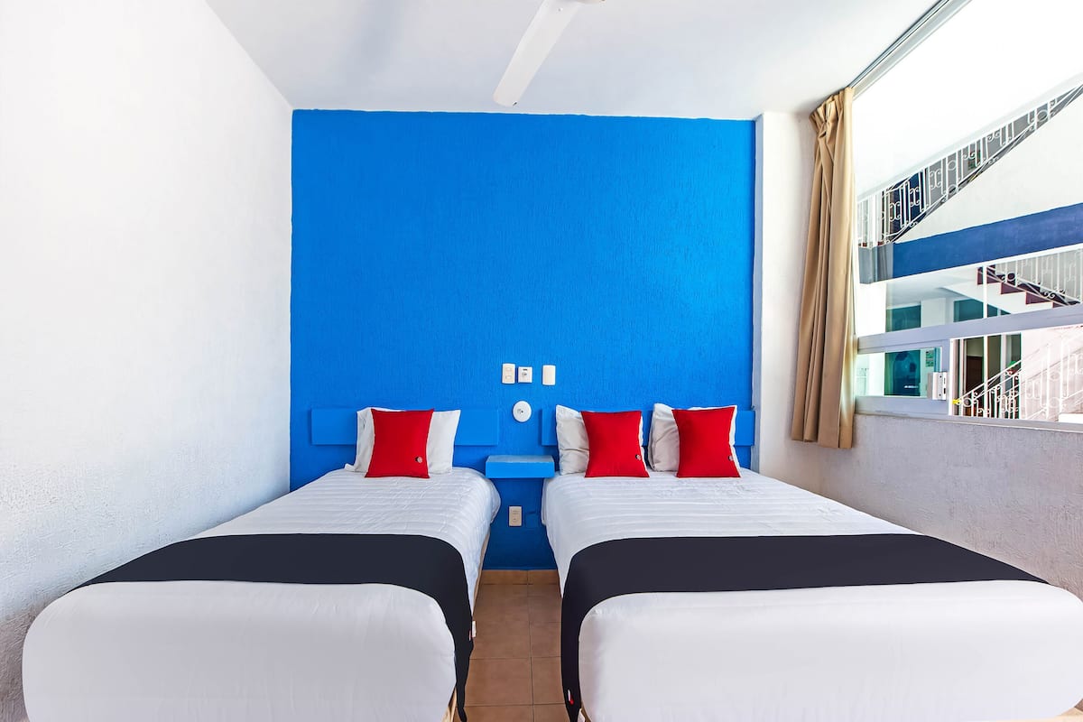 Habitación de hotel con dos camas, cada una con ropa de cama blanca y roja, una pared de acento azul y una ventana con vistas a una escalera.