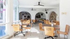 Una zona de menjador àmplia amb diverses taules i cadires, una cuina al fons i vistes a edificis a través de grans finestres.