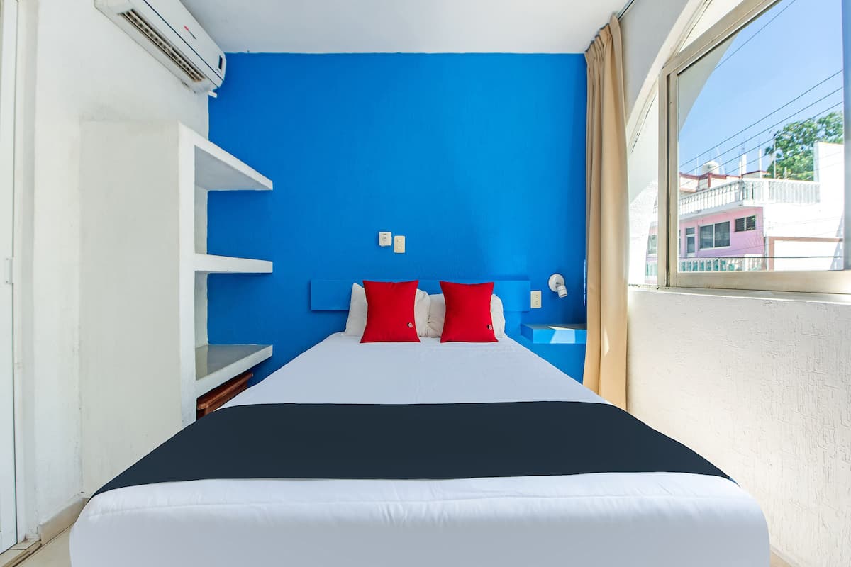 Habitación de hotel con cama, paredes blancas, una pared de acento azul y una ventana con vistas a edificios.