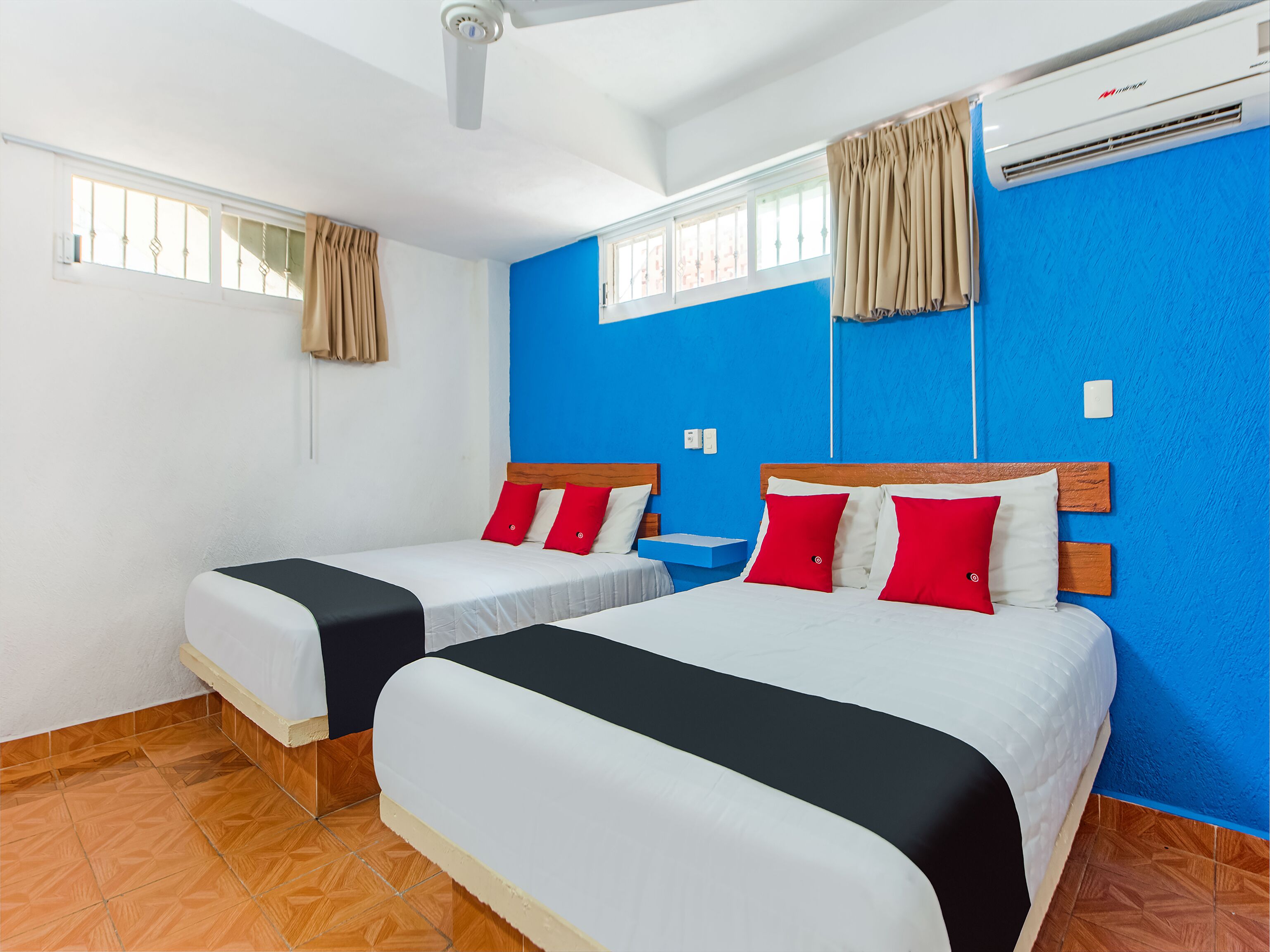 A hotel room with two beds, a blue accent wall, a large window with curtains, and an air conditioning unit.