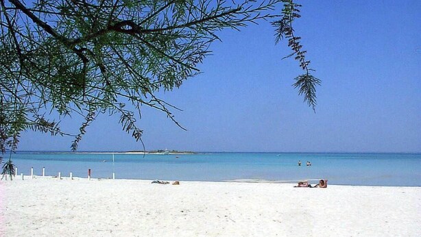 Une plage avec une eau d’un bleu limpide, du sable blanc et quelques personnes.