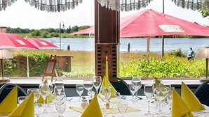 A table set for dining with a view of a body of water, red umbrellas, and a person walking in the background. - Hotel Elbblick (Geesthacht)