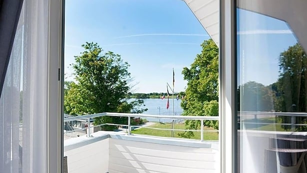 A balcony with a view of a lake, white railings, and a wicker chair. - Hotel Elbblick (Geesthacht)