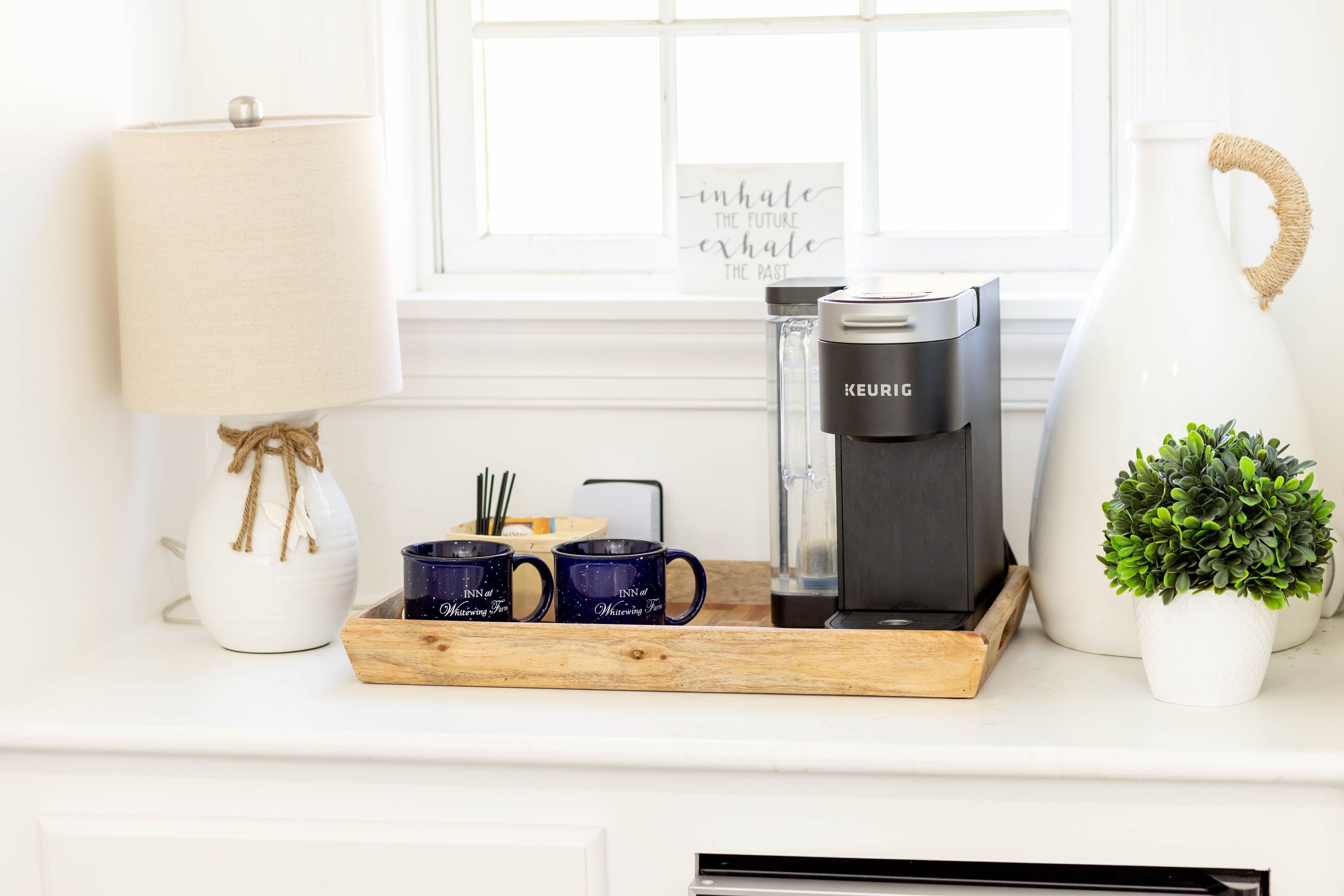 Una encimera de cocina con una cafetera, dos tazas, una lámpara y una planta en maceta.