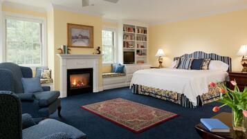 A bedroom with a fireplace, a large bed, a bookshelf, and a window with a view of trees.