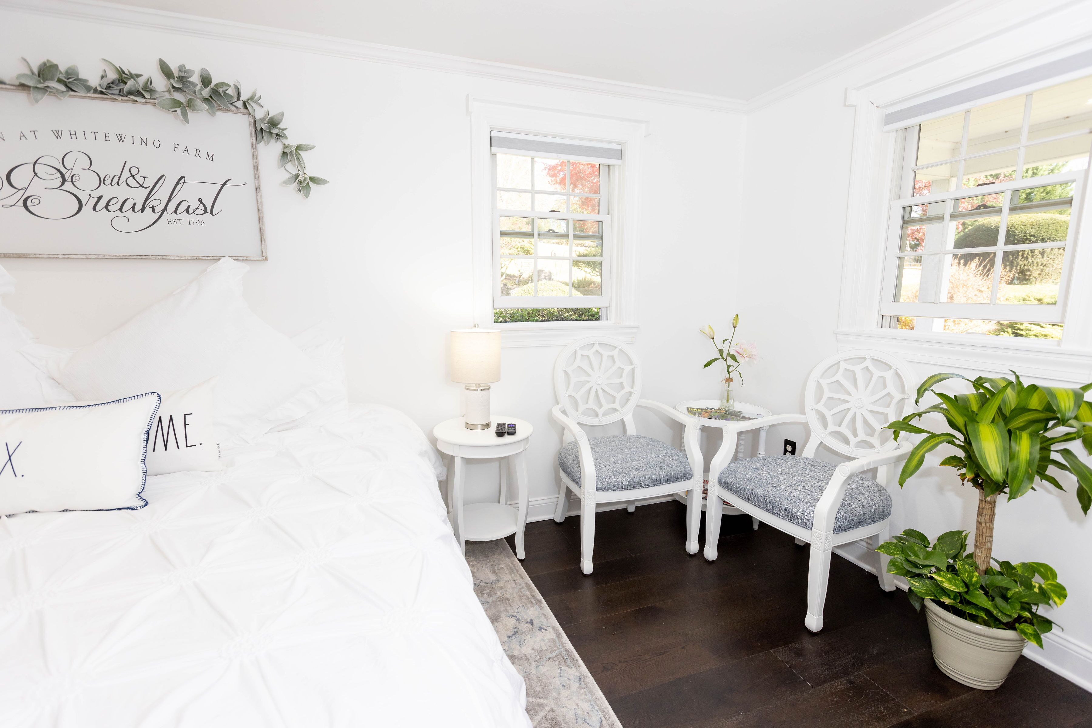 A white room with a bed, two chairs, a small table with a lamp, and a potted plant.