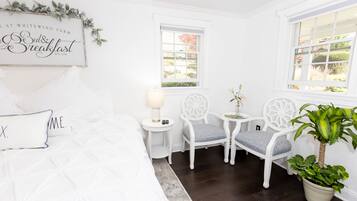 A white room with a bed, two chairs, a small table with a lamp, and a potted plant.