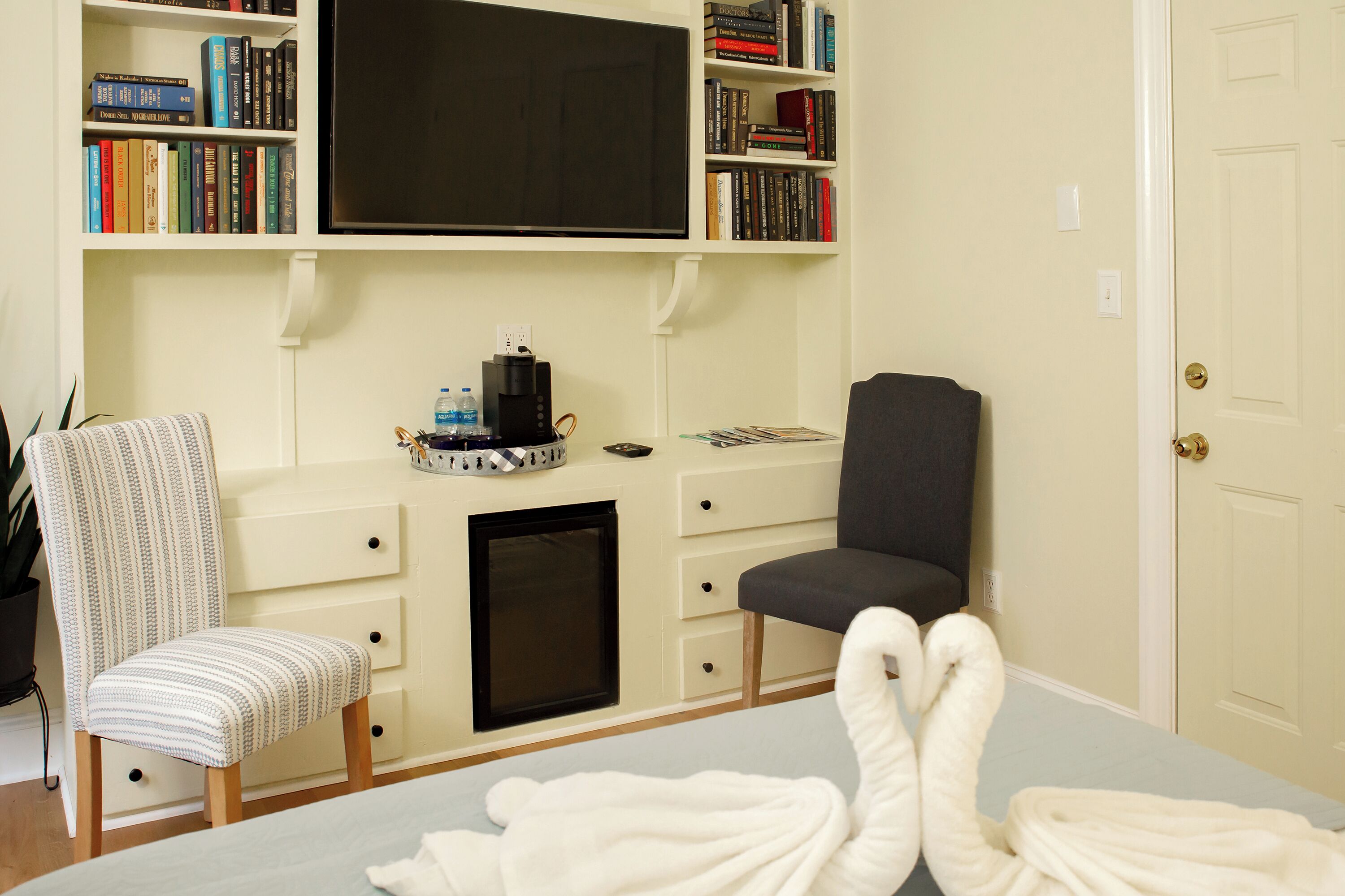 A hotel room with a bed, a desk with a computer, a chair, a bookshelf with books, and a television.