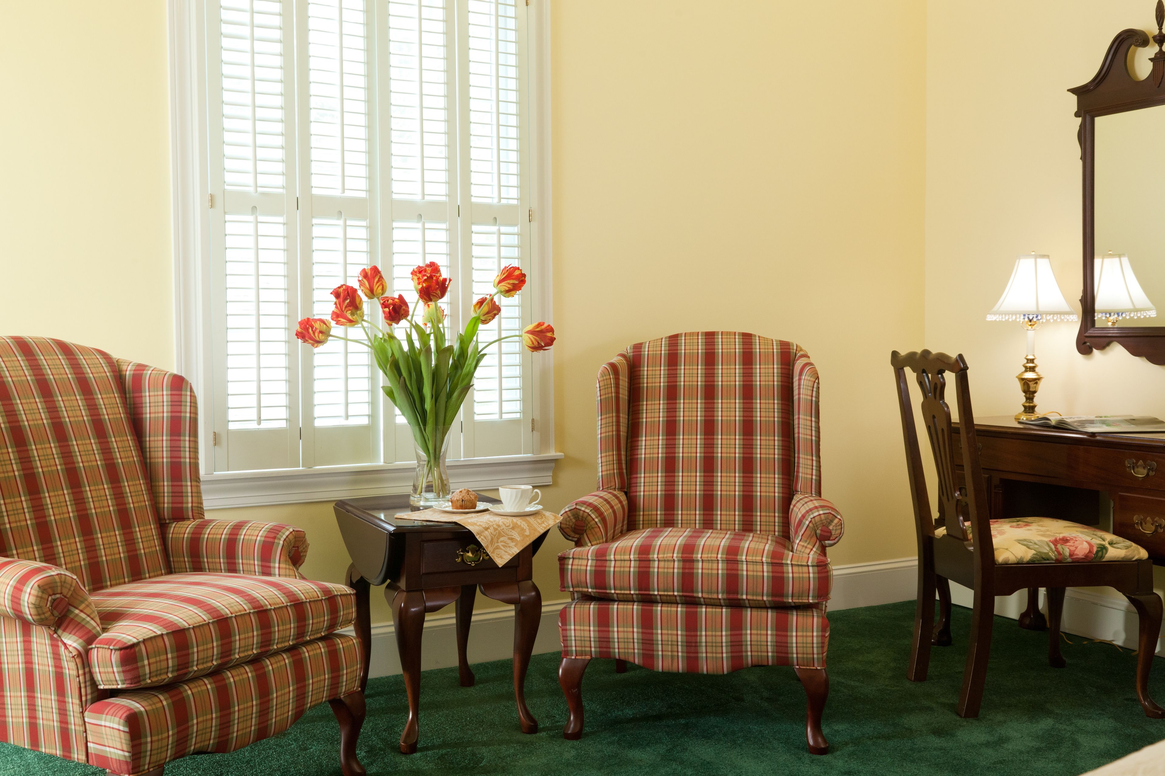 Ein Zimmer mit zwei karierten Sesseln, einem Holztisch mit Stuhl, einer Vase mit Blumen und einem Fenster mit Fensterläden.