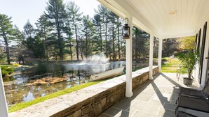 A covered patio with stone walls, a pond with a boat, and a forested area in the background. - The Inn at Whitewing Farm (West Chester)