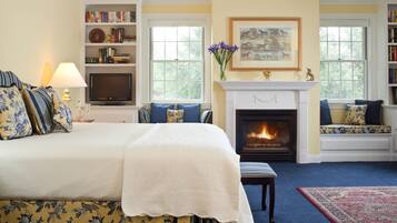 A bedroom with a large bed, a fireplace, a bookshelf, and a window with a view of trees.