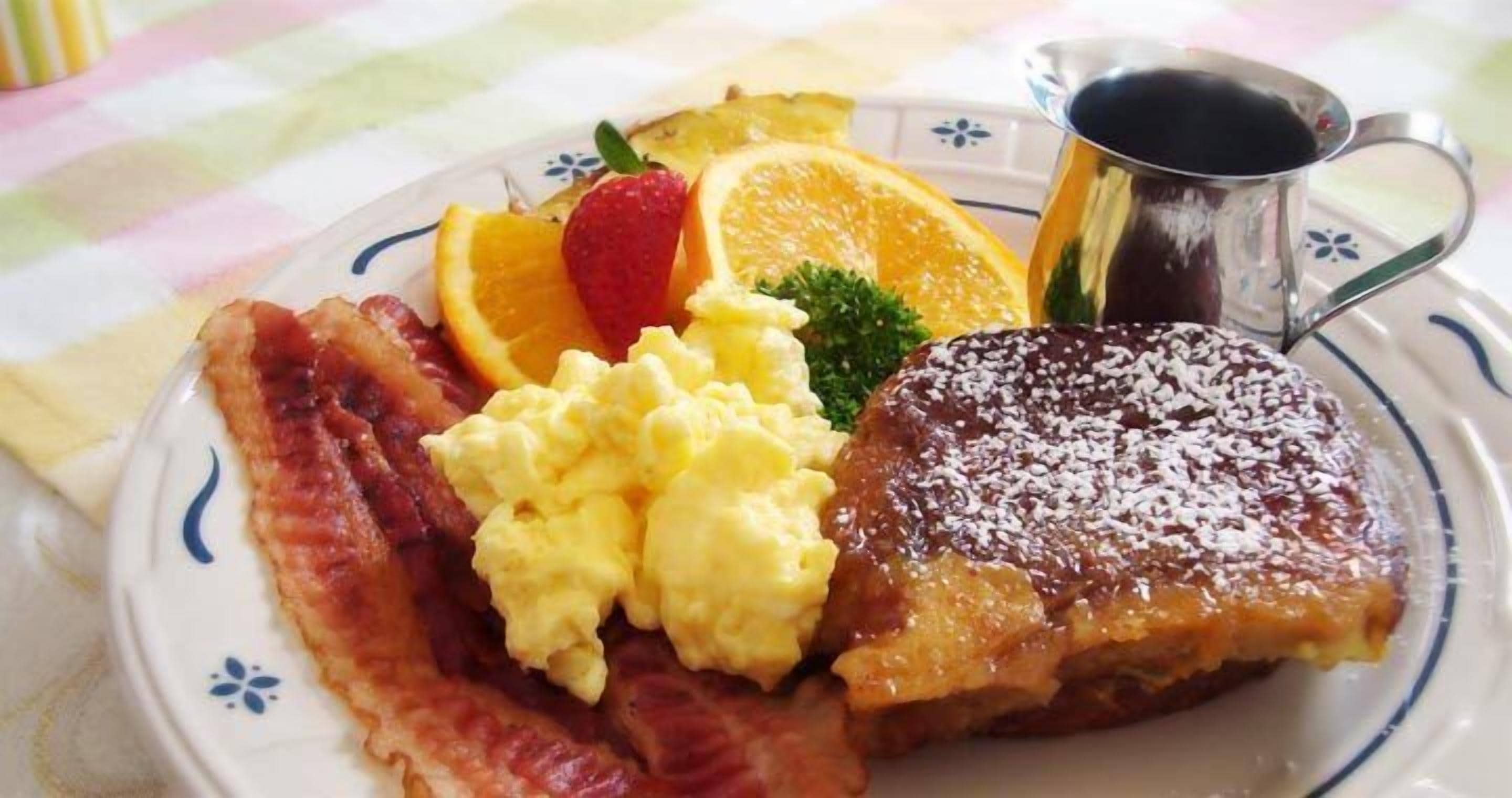 A plate with scrambled eggs, bacon, toast, orange slices, a strawberry, and a cup of coffee.