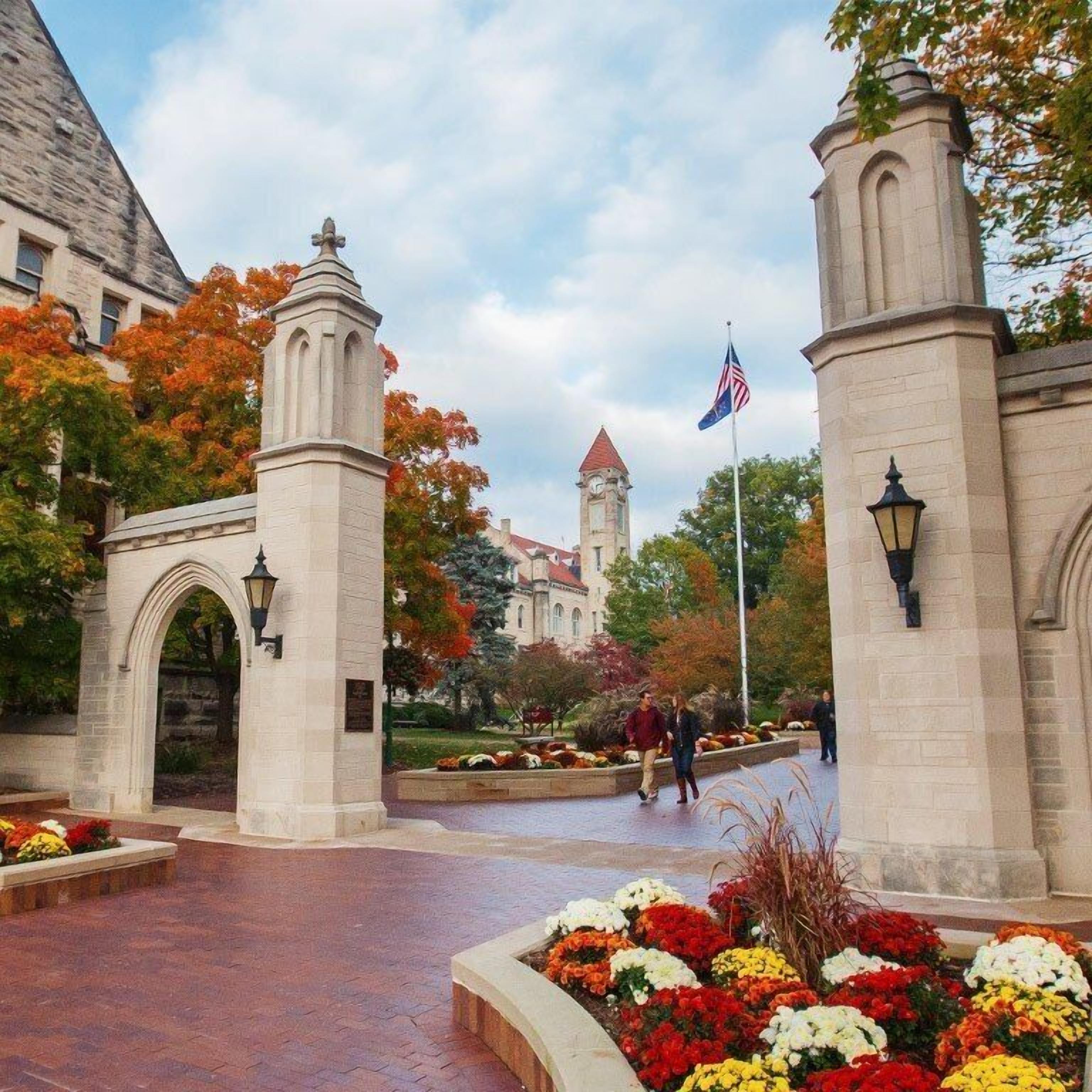 A historic building entrance with a clock tower, a flag, and a paved walkway with flower beds.