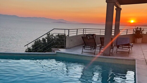 Un espace aménagé au bord de la piscine, offrant une vue sur le coucher de soleil en mer, avec une pergola et des sièges d’extérieur.