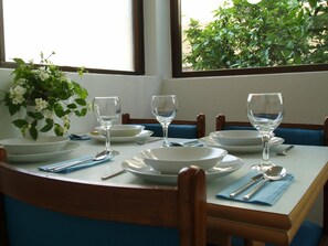 Una mesa de comedor dispuesta con platos, cuencos y copas de vino blancos, con un jarrón de flores blancas a la izquierda.