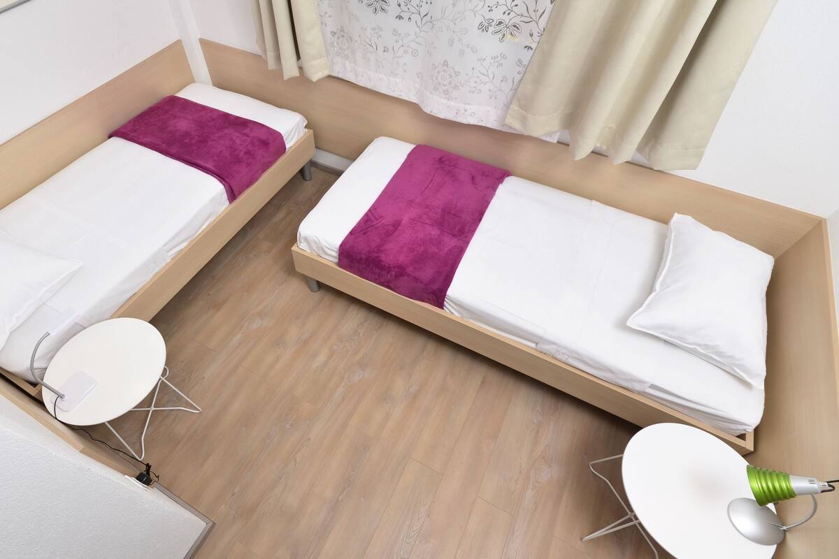 A room with two single beds, each with a white mattress and a purple blanket. There is a small round table with a white surface and a green lamp on the right side.