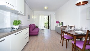Cozinha moderna com armários brancos, bancada de madeira e um vaso com planta. Há um sofá roxo e uma mesa de jantar com cadeiras na sala de estar.