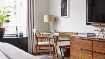 Une chambre d’hôtel avec un lit, un bureau avec une lampe, une chaise, une télévision, une bibliothèque et une fenêtre avec une plante.
