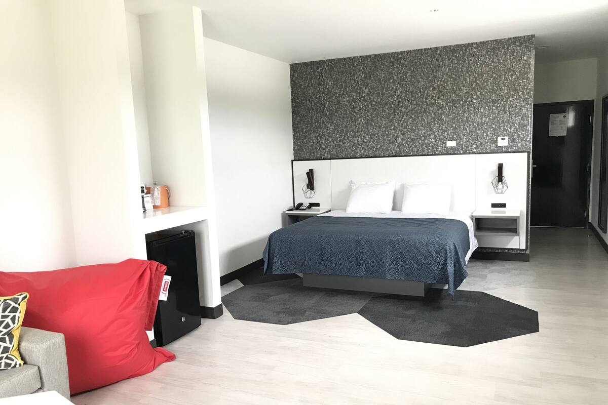 A modern hotel room with a bed, a red couch, a small table, and a black storage unit.