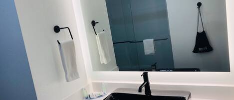 A bathroom with a white sink, black faucet, and a mirror reflecting a towel rack and a black bag.