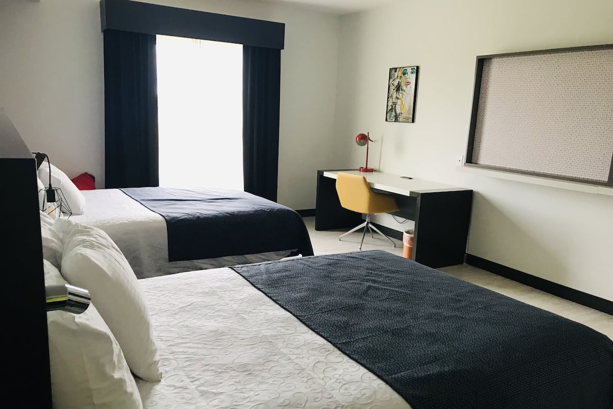 A hotel room with two beds, a desk, a chair, a large window with curtains, and a wall-mounted screen.