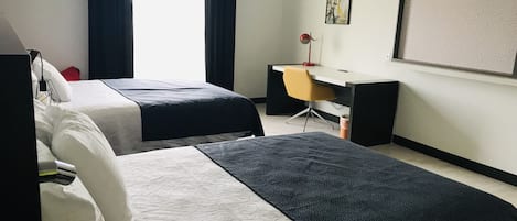 Habitación de hotel con dos camas, un escritorio, una silla, un ventanal con cortinas y una pantalla montada en la pared.