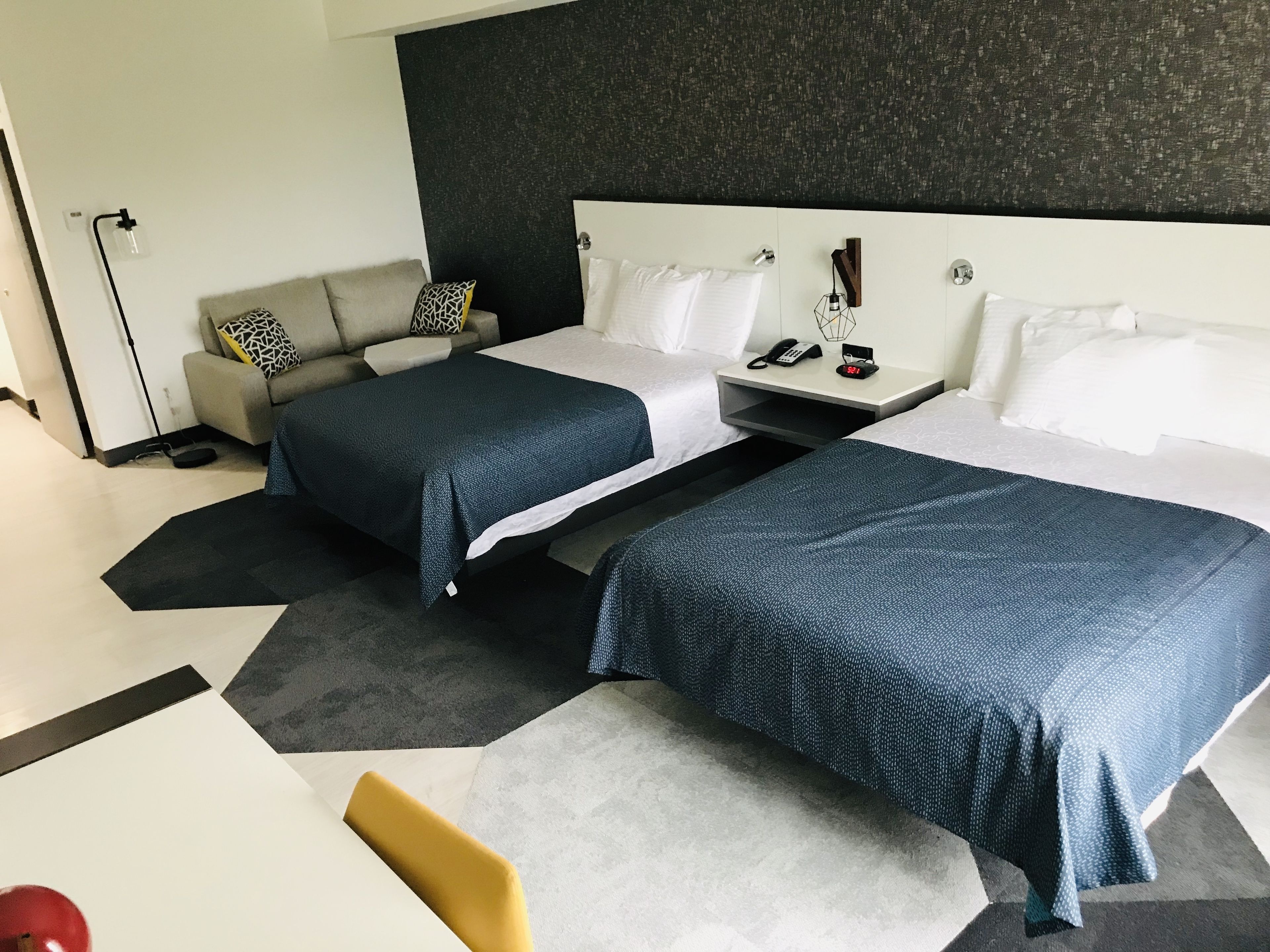 Ett modernt hotellrum med två sängar, en soffa, ett skrivbord och ett sängbord med en telefon och en väckarklocka.