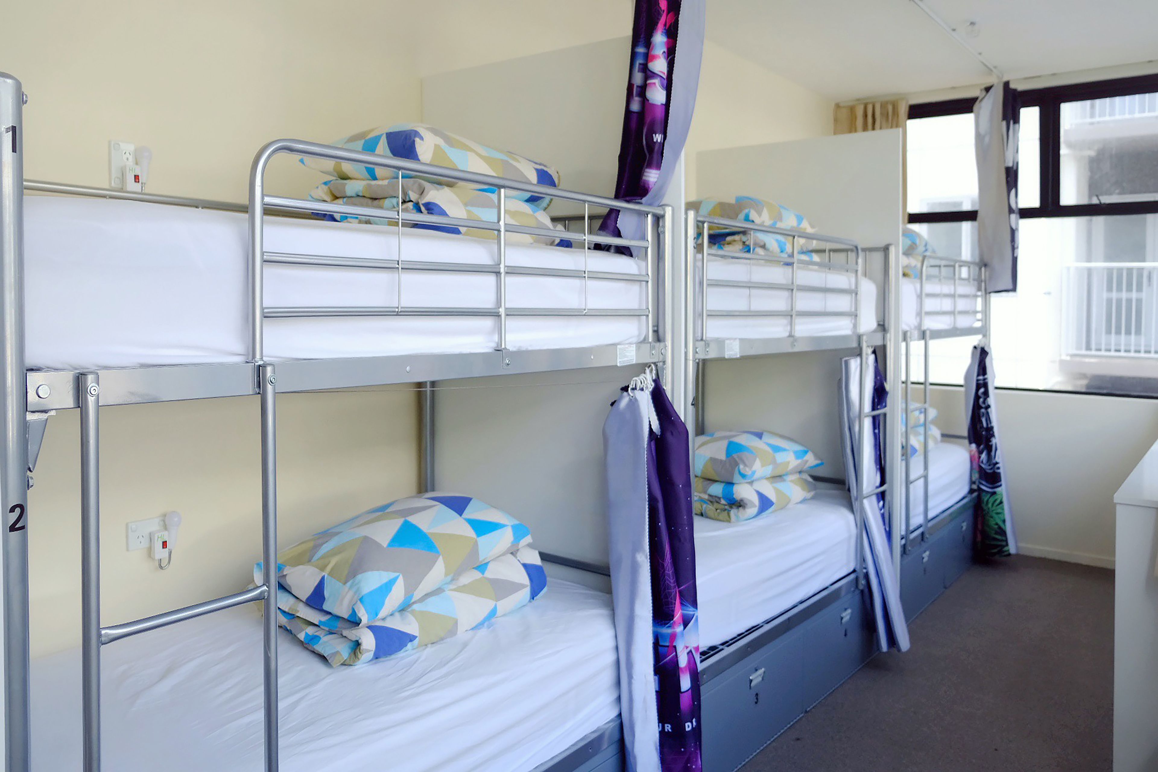 Une chambre avec un lit superposé, comportant deux lits, chacun avec une literie à motifs. On y trouve un petit bureau avec une chaise et une fenêtre avec des rideaux.