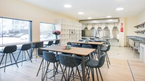 A modern restaurant with high tables and chairs, a large window, and a kitchen area. - Hobson Lodge - Hostel (Auckland)
