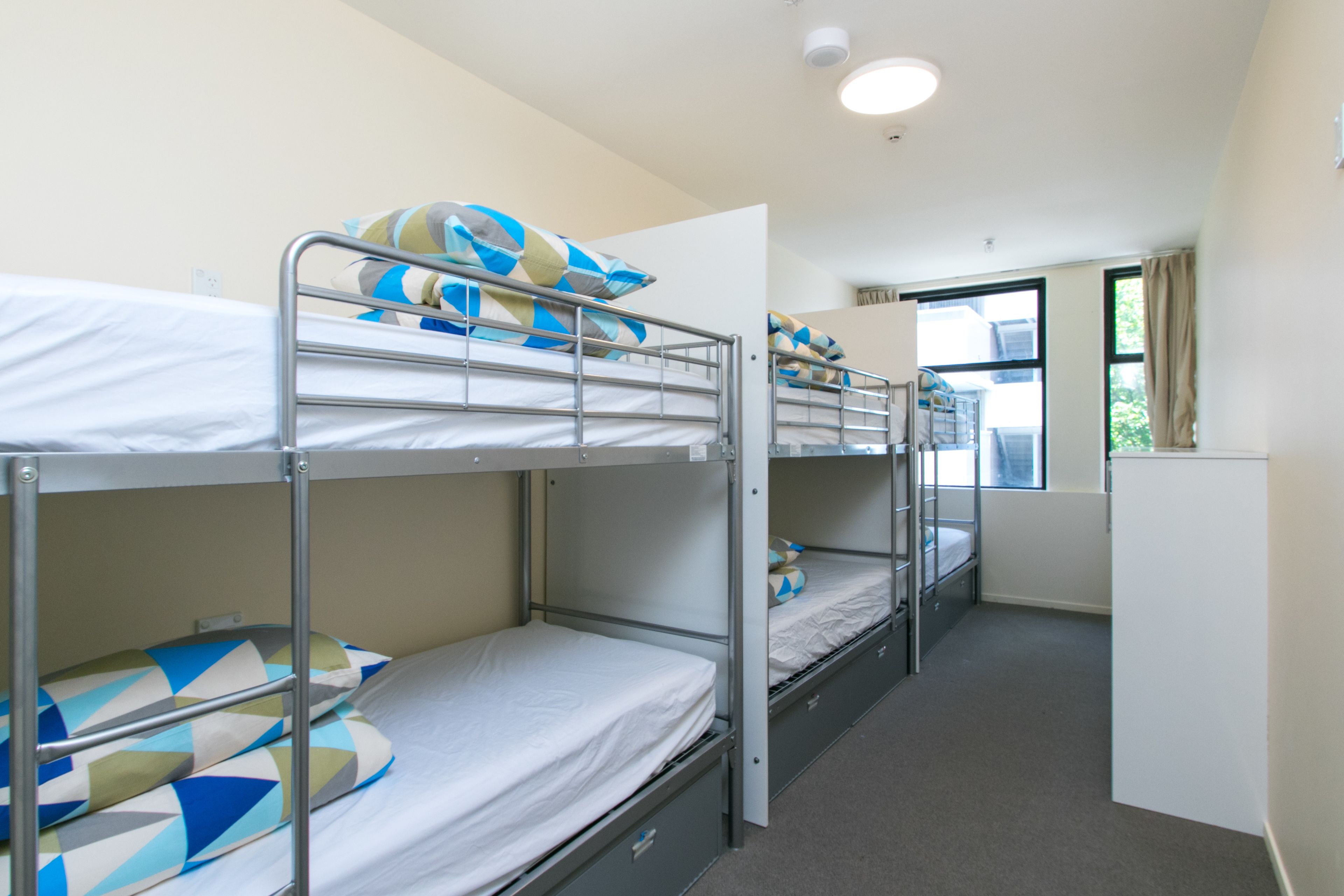 A dormitory room with bunk beds, a window with curtains, and a white wall.