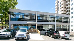 A modern building with a glass facade, a sign for Hobson Lodge, and several parked vehicles in front. - Hobson Lodge - Hostel (Auckland)