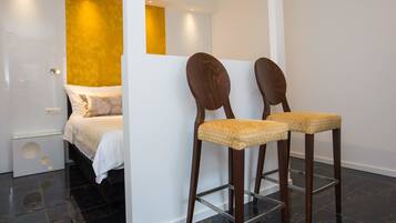 A modern hotel room with a bed, two wooden chairs with woven seats, a glass side table, and a yellow accent wall.
