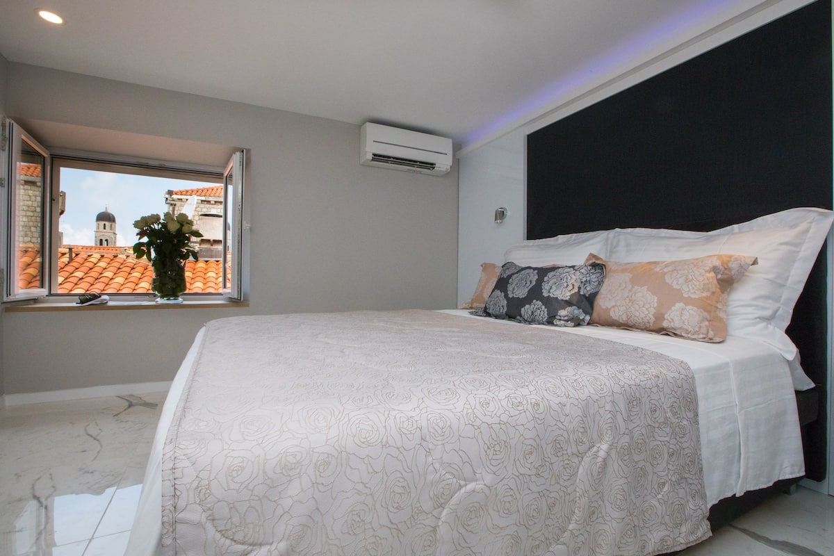 A bedroom with a large bed, a window with a view of buildings, an air conditioning unit, and a dark headboard.