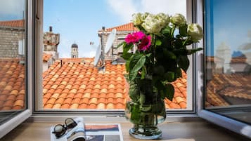 Una ventana con vistas a tejados de tejas color terracota, un edificio blanco con una cúpula, y un florero con flores en el alféizar.