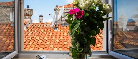 Una ventana con vistas a tejados de tejas color terracota, un edificio blanco con una cúpula, y un florero con flores en el alféizar.