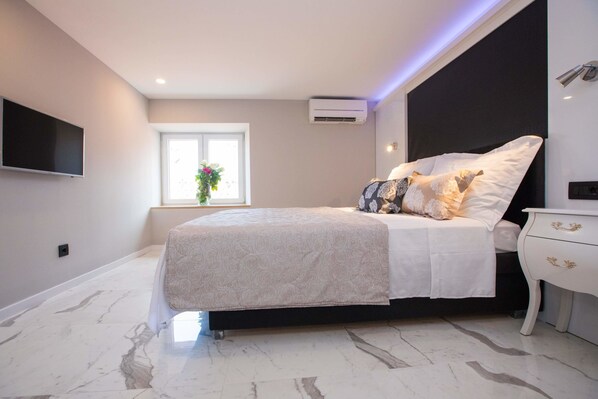 A modern bedroom with a large bed, a bedside table, a TV mounted on the wall, and an air conditioning unit.