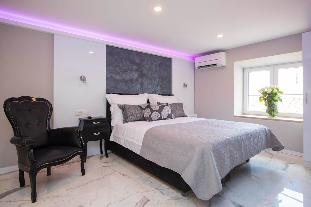 A bedroom with a bed, a nightstand, a chair, a window with a view, and an air conditioning unit.