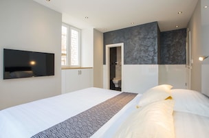 A modern bedroom with a large bed, a flat-screen TV mounted on the wall, a window, and a bathroom visible through an open door.