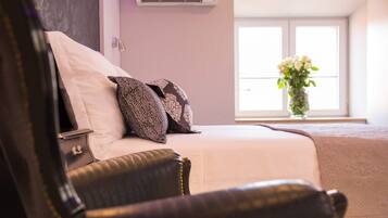 Een slaapkamer met een bed, kussens, een nachtkastje, een stoel en een raam met een bloevas.