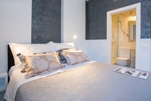 Cama bem arrumada com colcha estampada e dois travesseiros decorativos, mesa de cabeceira branca, e um banheiro visível por uma porta aberta.