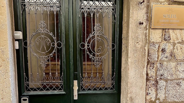 Puertas dobles verdes con celosías metálicas ornamentales, una pequeña placa en la parte superior y una pared de piedra a la derecha.